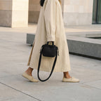 Woman walking outdoors holding a PLSH designer black handbag, wearing a beige coat and shoes.