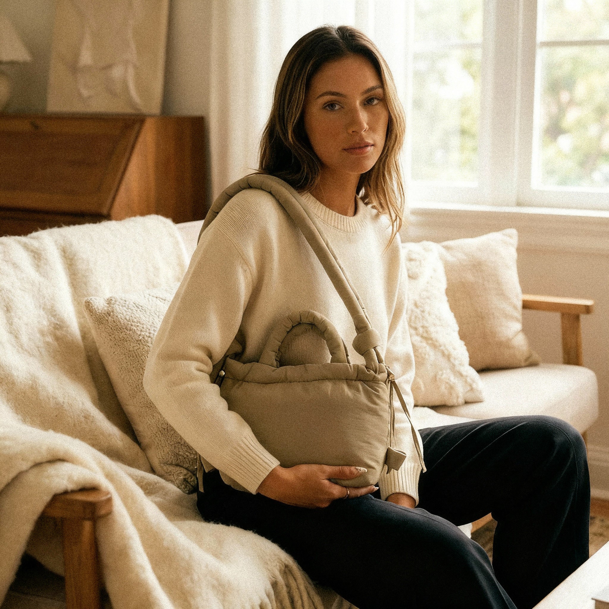 Woman sitting on a couch holding a PLSH designer beige handbag, wearing a white sweater.