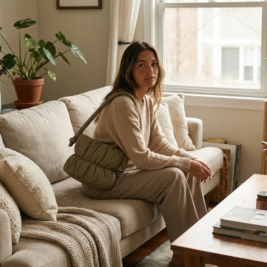 Woman sitting on a couch wearing a PLSH designer handbag in a cozy living room with a plant and books on a table