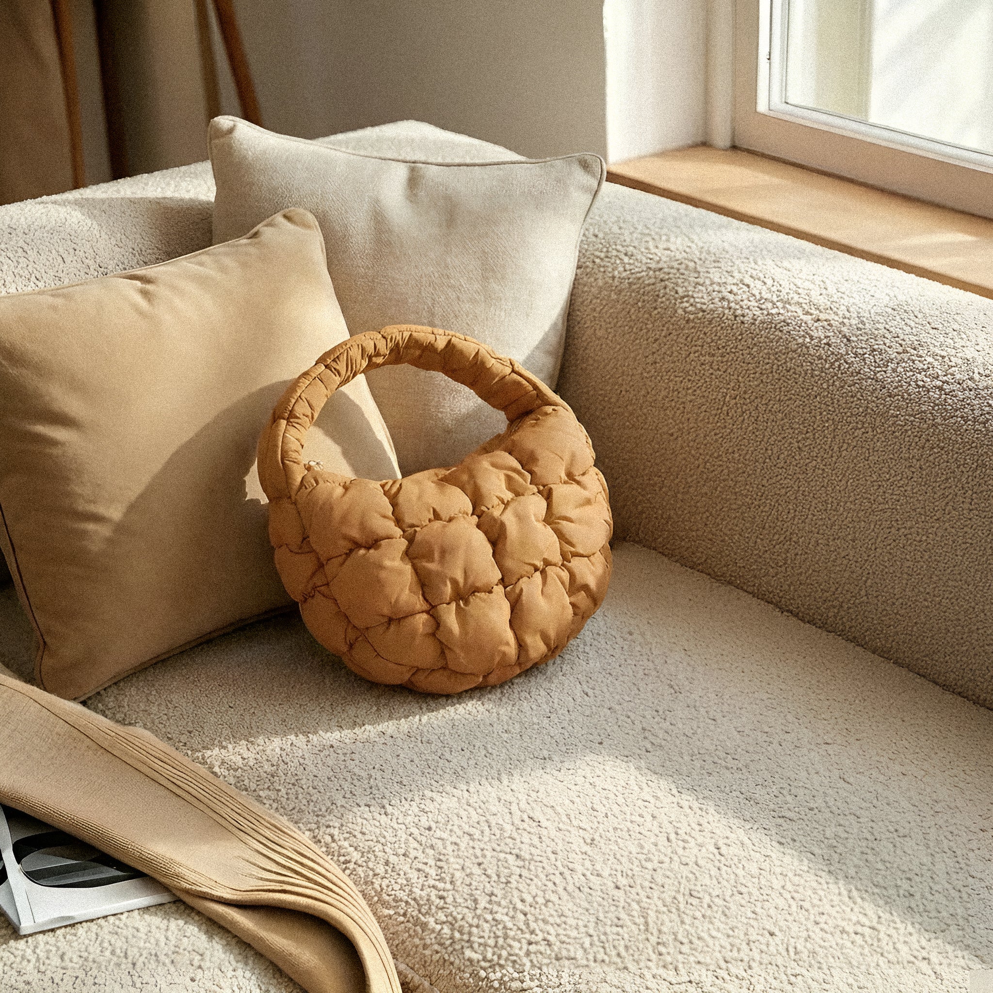 PLSH brown women's designer quilted handbag on a beige sofa with pillows and a window in the background