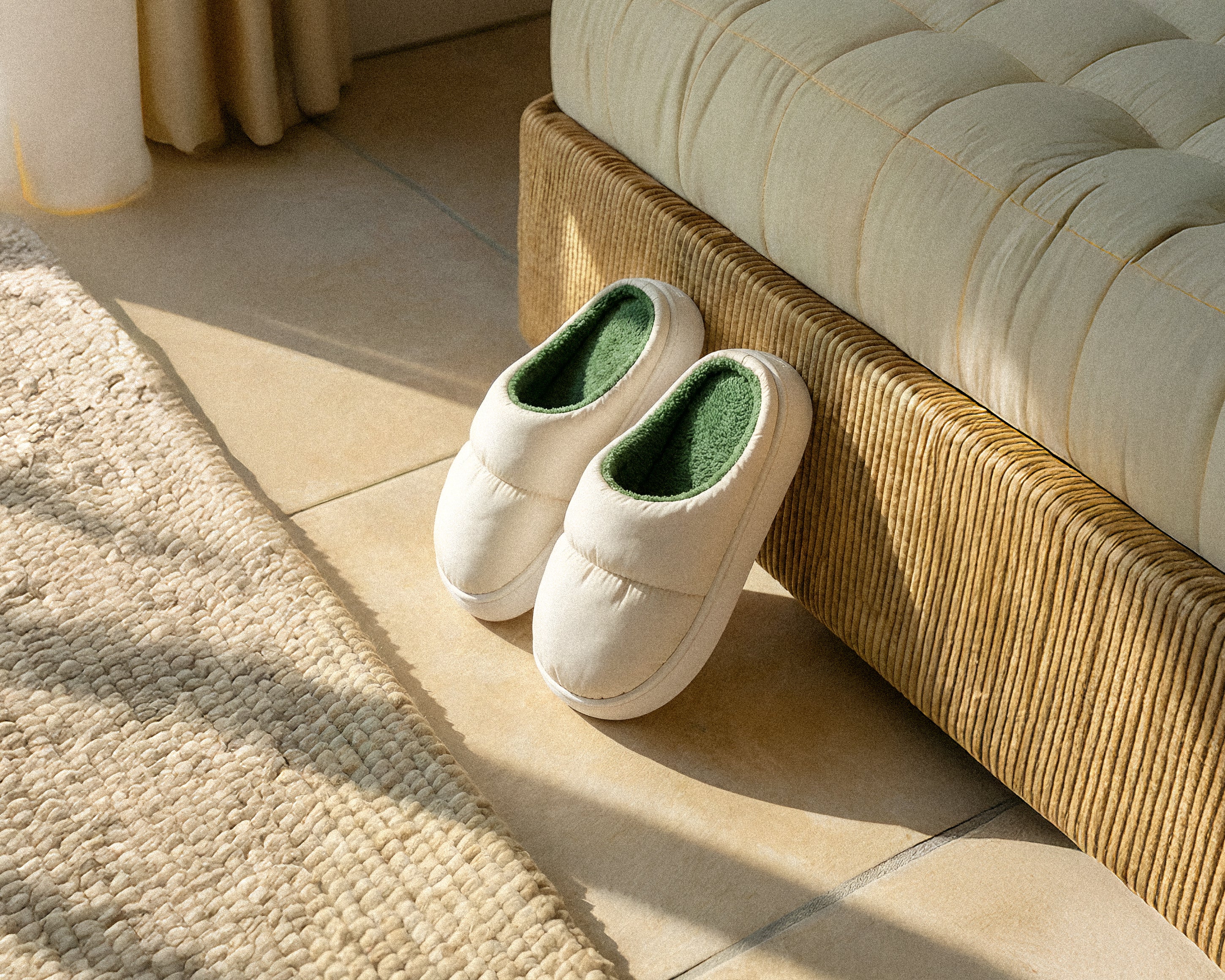 Pair of white PLSH premium slippers with green interior against a bed next to a beige carpet